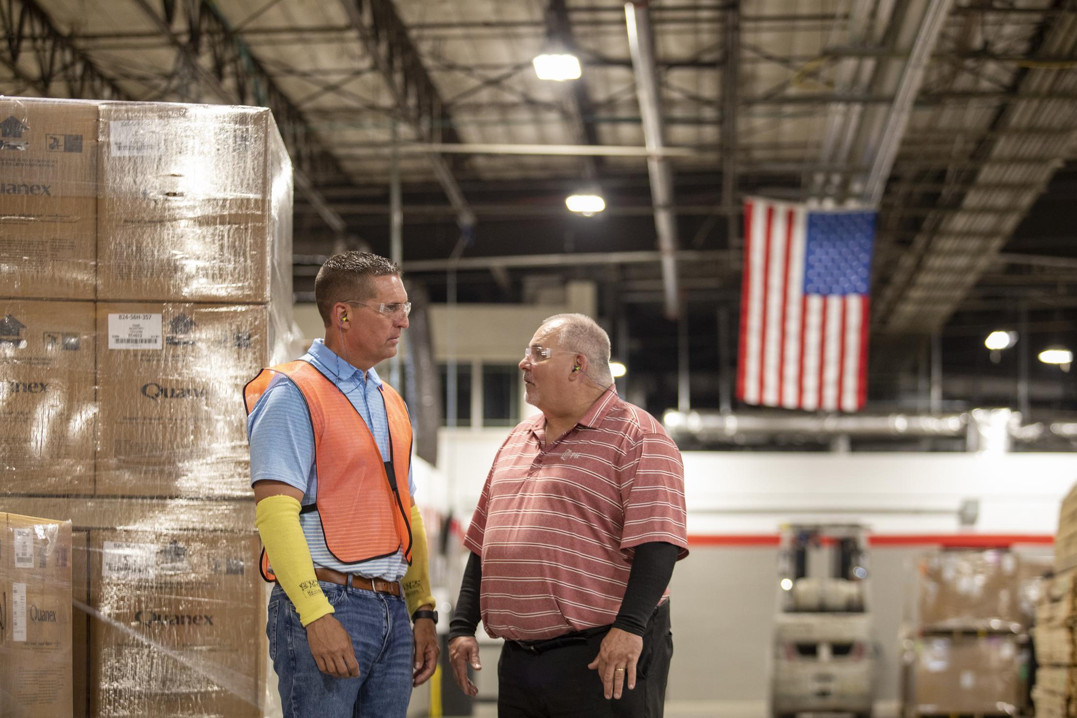 Two men talking in a manufacturing plant with the United States of America flag hanging in the background.