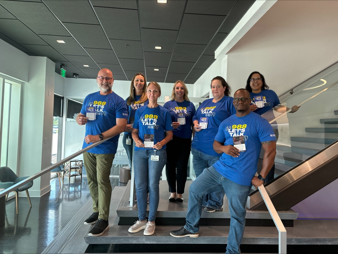 From left to right: MITER Brands team members: Bryan Lingle, Jennifer Meyer, Amy Lowery, Amie Brooks-Huff, Coley Pelletier, Ray Jones, and Veronica White wearing their “988 Let’s Talk” t-shirts and displaying information cards in honor of National Suicide Prevention Week